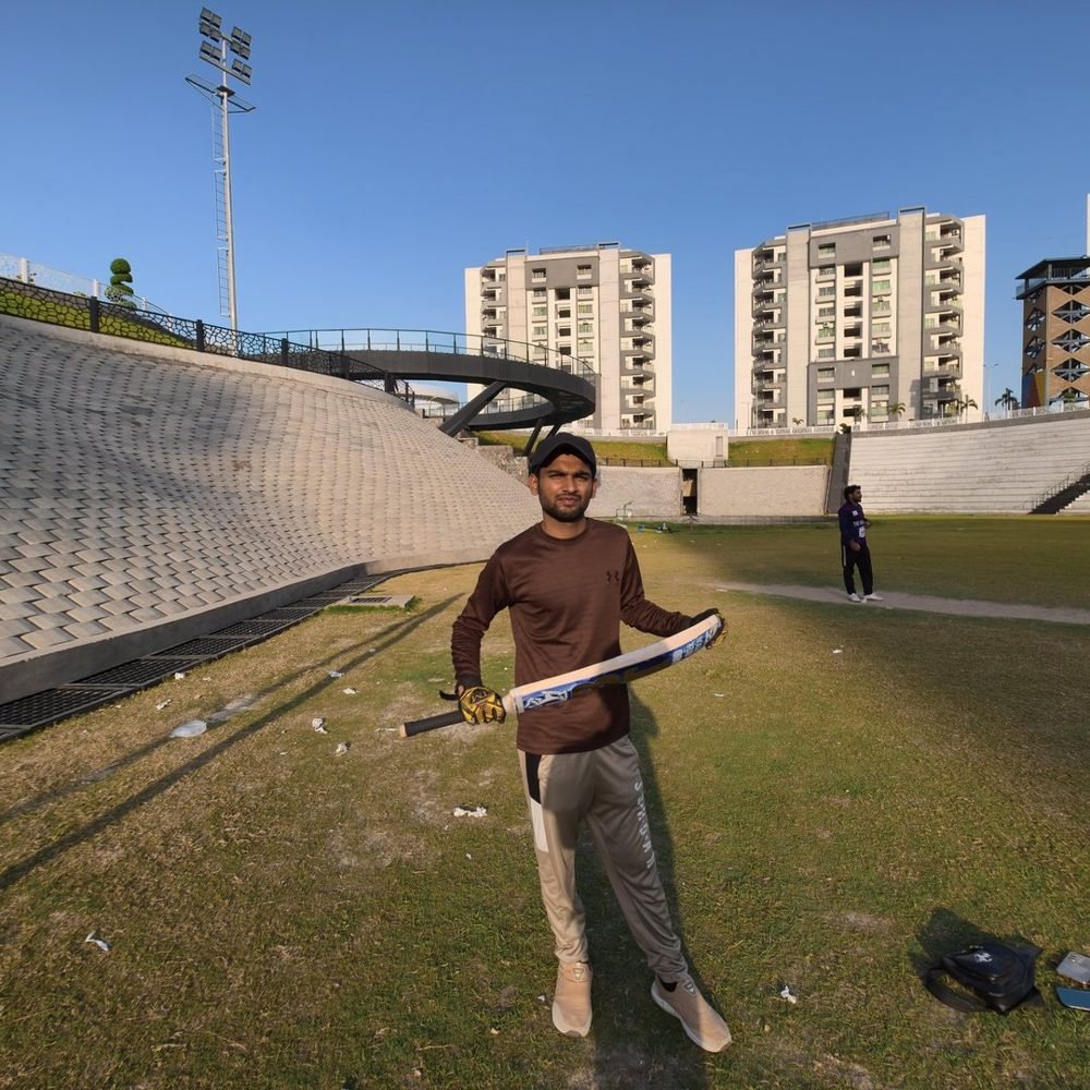 Waleed holding a cricket bat on a ground with apartment blocks behind